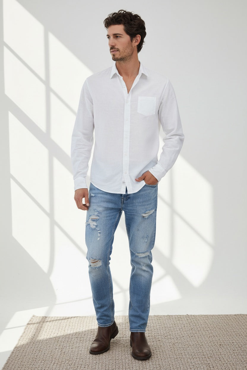 A man stands indoors by a window, wearing the FRANK & EILEEN DON Men’s Button Up Shirt in Classic Featherweight Cotton White, paired with light blue ripped jeans and brown boots. With one hand in his pocket and a neutral expression, sunlight casts shadows on the white wall behind him, highlighting the effortless appeal of this everyday essential from FRANK & EILEEN.