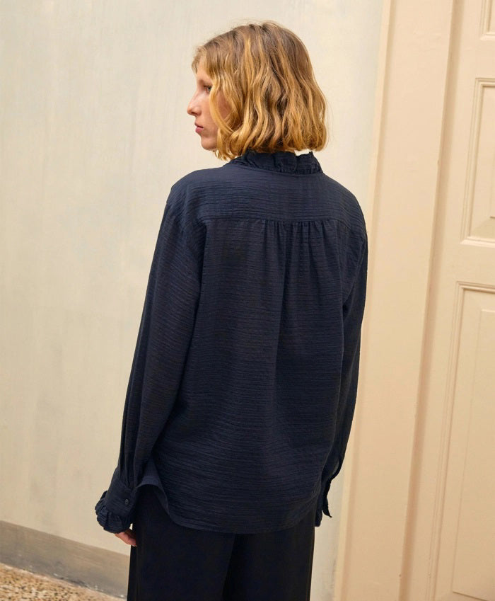 A person with shoulder-length blonde hair is wearing the MOMONI - Marc Blouse in textured cotton silk, which features ruffles along the collar and cuffs. They are also wearing black wide-leg pants and holding a teal-colored clutch. The background includes a light-colored door and wall.