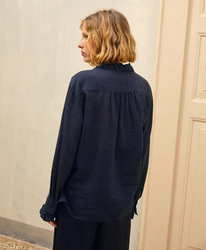 A person with shoulder-length blonde hair is wearing the MOMONI - Marc Blouse in textured cotton silk, which features ruffles along the collar and cuffs. They are also wearing black wide-leg pants and holding a teal-colored clutch. The background includes a light-colored door and wall.