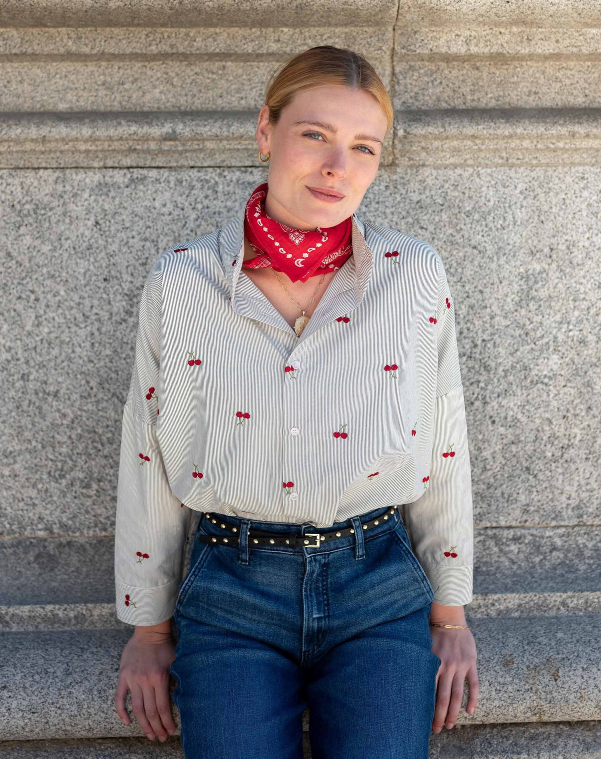 A woman with blonde hair in a ponytail, dressed in the MERSEA Emerson Button Up Shirt in Cherries On Top by MERSEA and blue jeans, holds a red clutch while standing outdoors near green trees and patterned tiles, smiling and looking to the side.