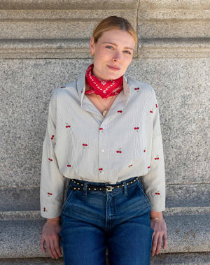 A woman with blonde hair in a ponytail, dressed in the MERSEA Emerson Button Up Shirt in Cherries On Top by MERSEA and blue jeans, holds a red clutch while standing outdoors near green trees and patterned tiles, smiling and looking to the side.