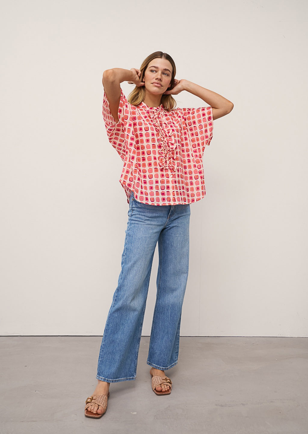 A woman wears the DAISY BLOUSE IN PINK PRINT by 0039 ITALY, featuring pink and red square patterns with ruffles, paired with light blue jeans against a plain light background.
