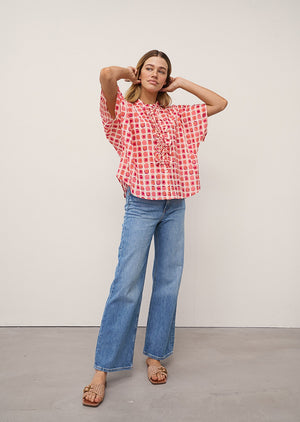 A woman wears the DAISY BLOUSE IN PINK PRINT by 0039 ITALY, featuring pink and red square patterns with ruffles, paired with light blue jeans against a plain light background.