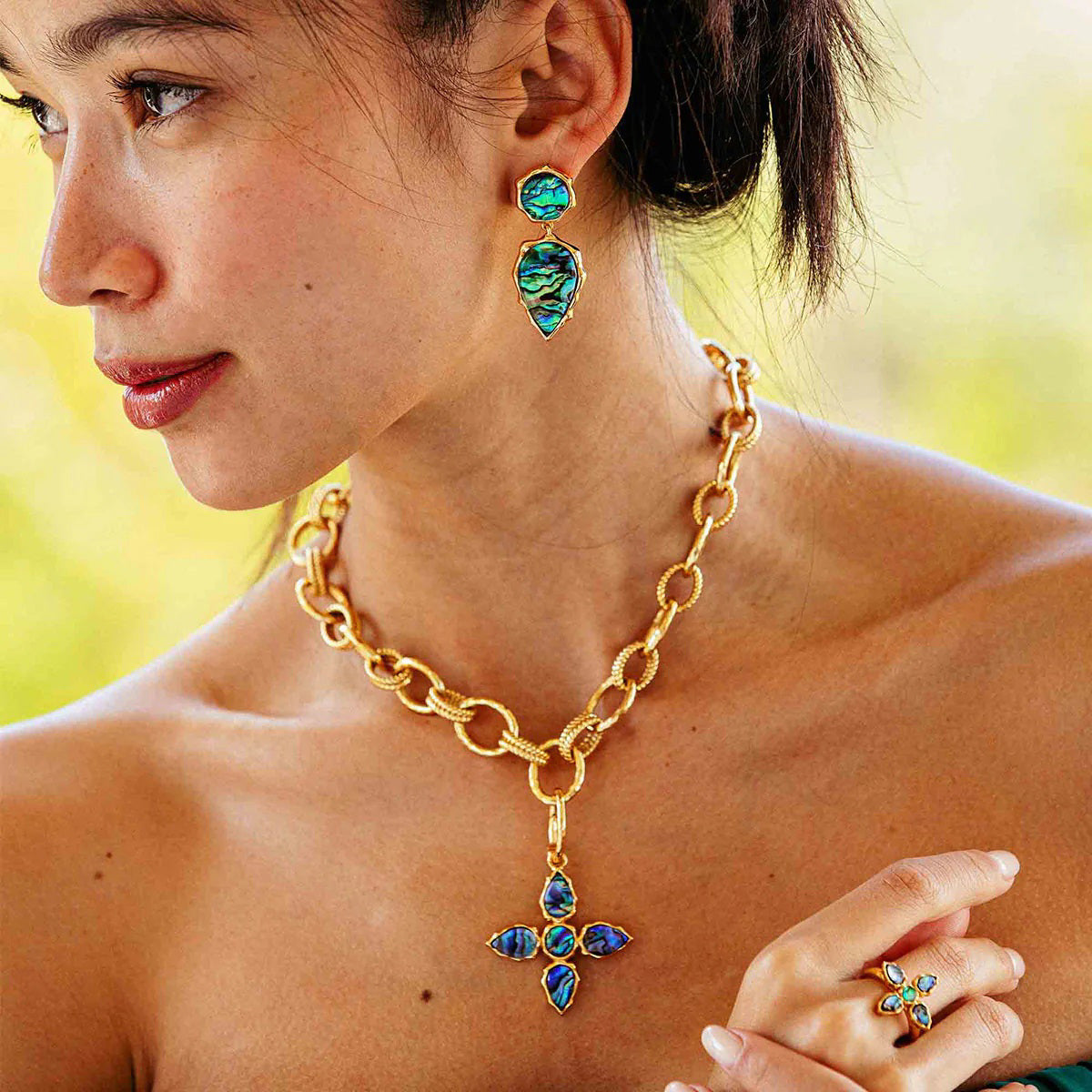 The CAPUCINE DE WULF AURORA pendant features a gold cross with four sustainably sourced blue-green abalone stones set in textured gold bezels. An elegant embellished loop enhances its wearability.