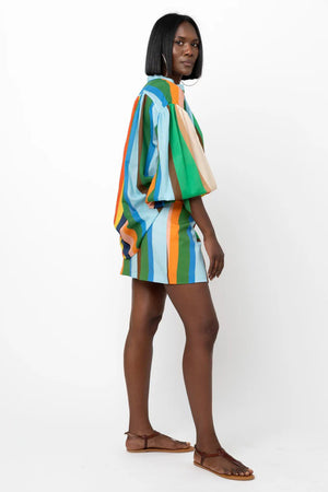 A person stands against a white background, wearing the HOPE FOR FLOWERS Wrist Tie Shirt in a blue and white geometric pattern, paired with a short, multicolored striped skirt. They have shoulder-length hair and are wearing sunglasses. Their hands rest by their sides.