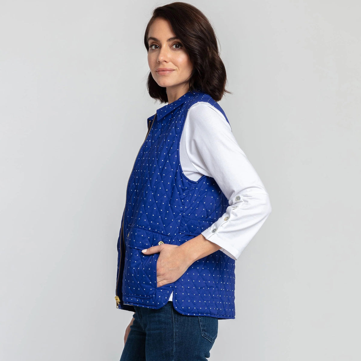 A woman with short brown hair is wearing the HINSON WU - LAUREN REVERSIBLE VEST in bright blue, paired with a white, long-sleeve blouse that features buttons on the cuffs. She completes her outfit with blue jeans and stands against a plain white background.