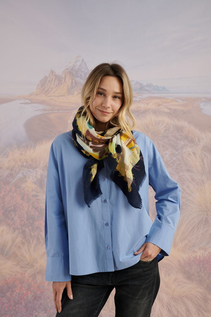 A woman with light brown hair wears a cream blouse and the STORIATIPIC - ANDRES WOOL SCARF by STORIATIPIC, elegantly hand-embroidered with green, yellow, red, and blue patterns. She gazes to the side against a scenic background.
