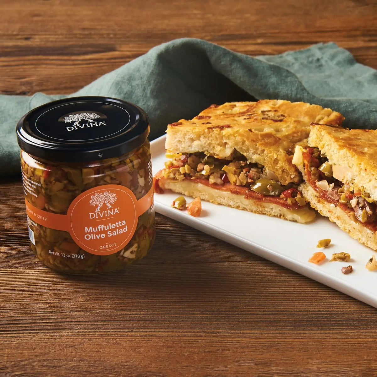 A clear glass jar of DIVINA - MUFFULETTA OLIVE SALAD from the brand DIVINA, inspired by the classic flavors made famous by Central Grocery in Greece. The orange label with product name and certifications, along with a black lid, highlights the authentic essence of traditional muffuletta olive salad.