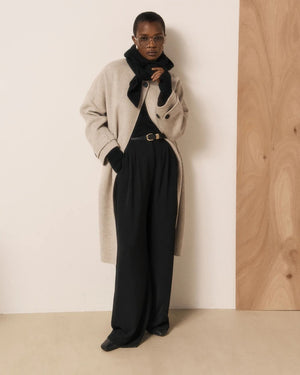 Wearing the HARTFORD - MERA Knit Hood/Scarf in Black—crafted from luxurious wool and cashmere—this person pairs a light long coat with a black turtleneck, wide-leg black pants, and gloves. They stand indoors against a light wall and wooden panel, striking a pose with one hand resting on their face.
