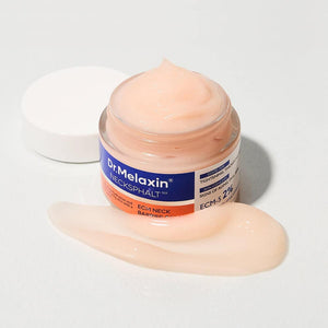 An open jar of KOSMIC KBEAUTY SKINCARE DR MELAXIN - NECKSPHALT ECM NECK BARRIER CREAM with light peach cream spread on the white surface in front of it.