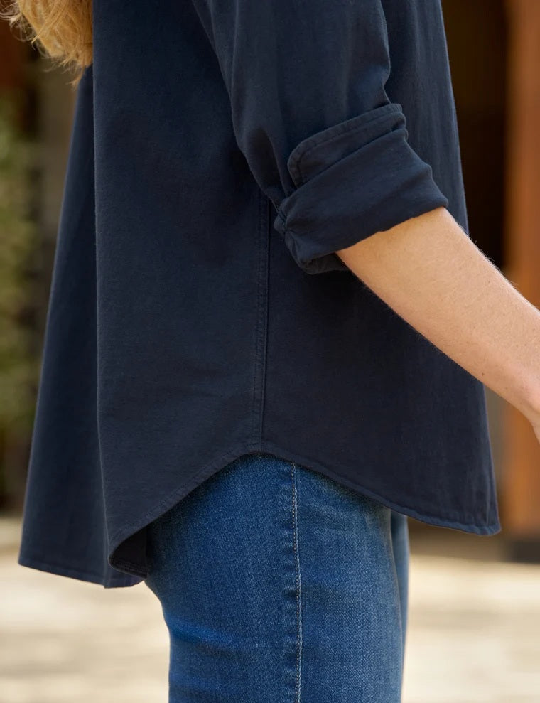 A woman stands in a minimal room, smiling with one hand in her pocket, wearing the FRANK & EILEEN - Waverley Classic Button Up Tee in Vintage Jersey Classic Navy from the California Heritage Collection, paired with blue jeans and brown ankle boots. A large window to the left fills the space with natural light.