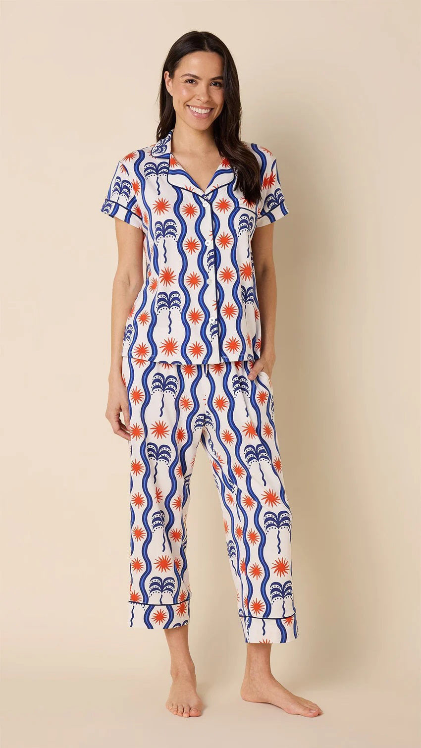 A person with long, dark hair stands barefoot on a beige background wearing CAT&#39;S PAJAMAS&#39; luxurious Red, White and Breeze Pima Knit Capri Set, featuring bold abstract designs.