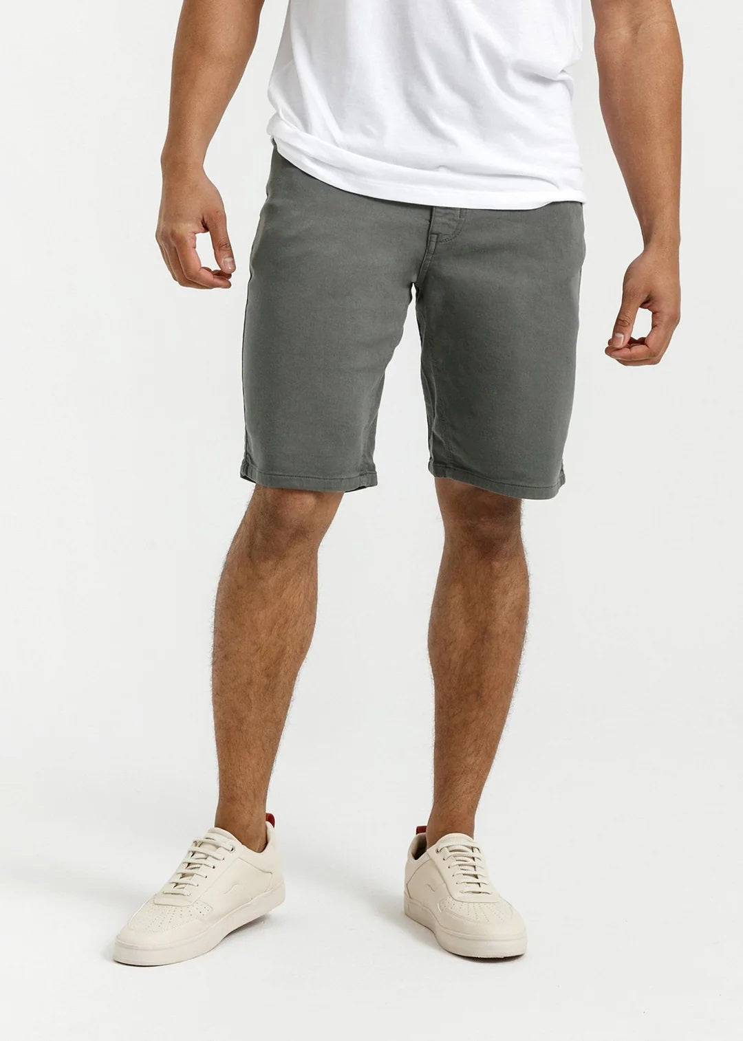 A person stands against a plain light background, wearing a white T-shirt, DUER No Sweat Relaxed Shorts in Gull made from stretch fabric, and white sneakers, with one hand in their pocket.