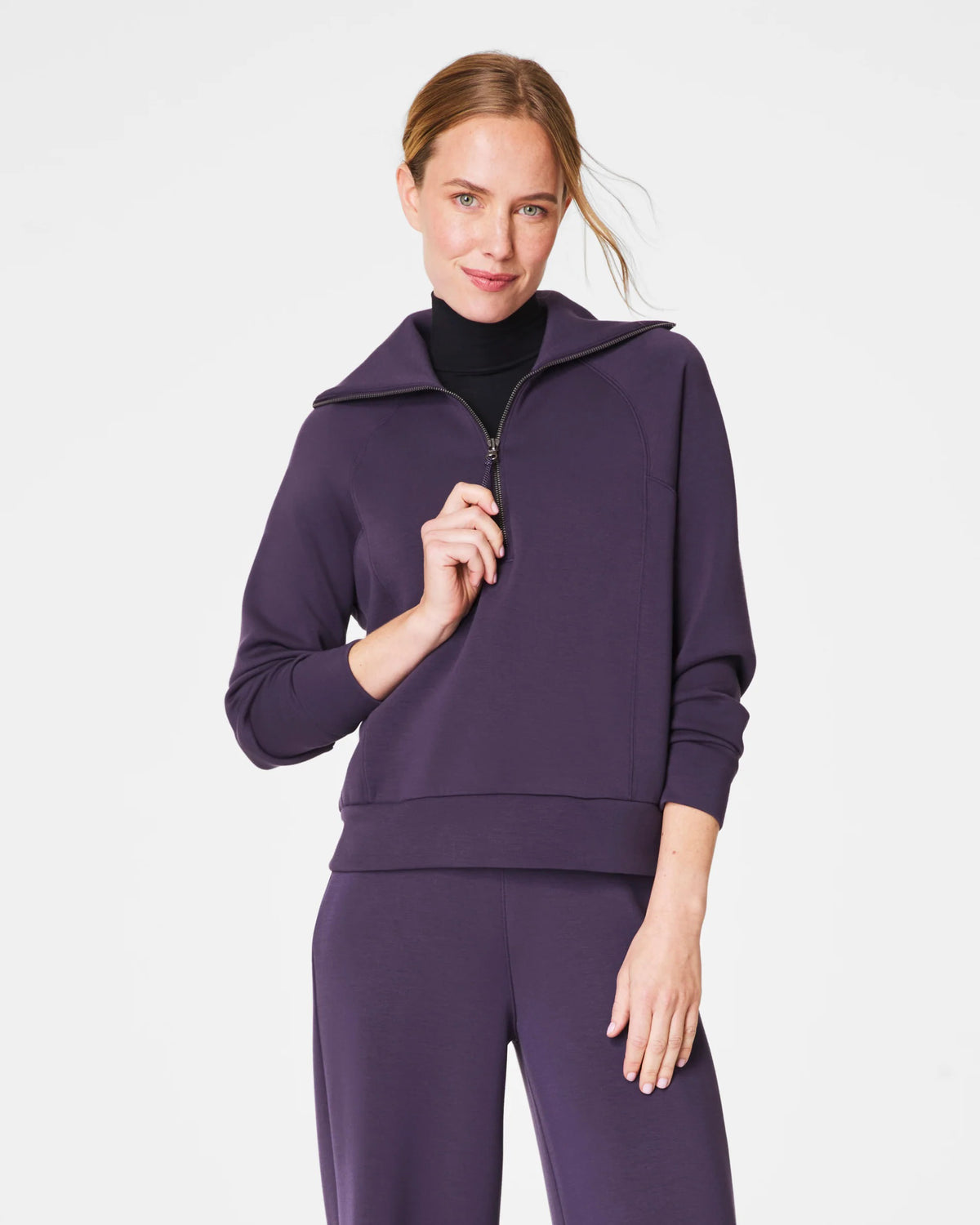 A person with short hair is wearing a SPANX AIRESSENTIALS HALF ZIP sweater in navy blue, made from lightweight spacer fabric, paired with black pants. Their arms are crossed over their chest as they stand against a plain, light grey background.