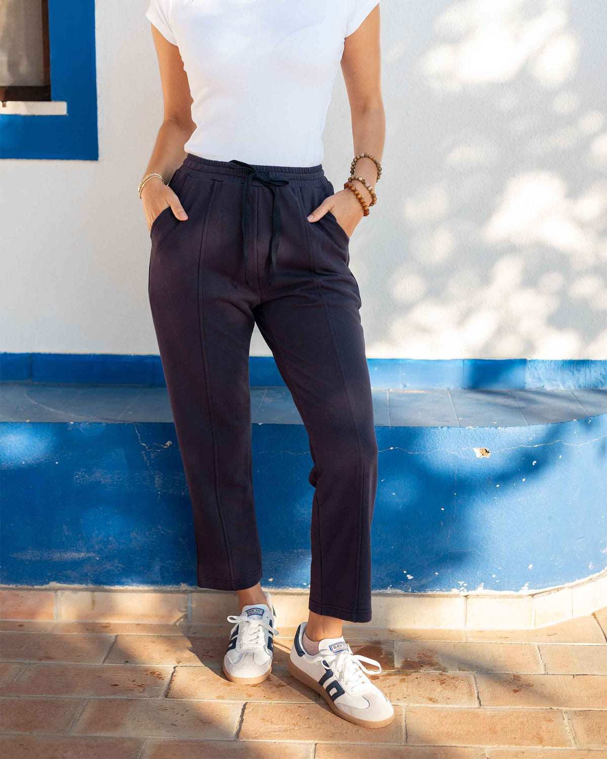 A person wears the MERSEA - FRENCHIE CIGARETTE PANT by MERSEA, paired with a white top and gray sneakers, standing on sandy beach ground with ocean waves and blue sky behind them, hands in their pockets.