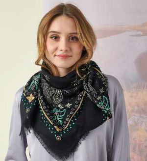 A young woman with light brown hair wears the STORIATIPIC Nelly Square Cashmere and Wool Embroidered Bandana Scarf, paired with a light gray top, standing before a softly colored nature-themed backdrop.