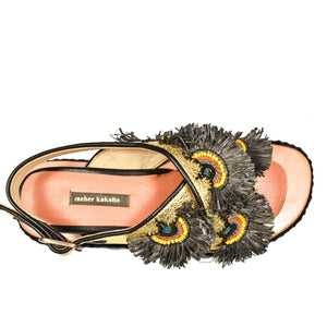 A person wearing MEHER KAKALIA's Rio Bloc Sandal in Charm Gold, featuring intricate, peacock feather-like designs and unmatched artisanal quality. With painted toenails and rolled-up jeans, they sit on a wooden floor.