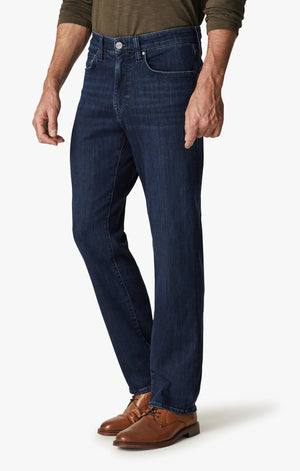 A man in a green long-sleeve shirt, brown shoes, and 34 HERITAGE Charisma Relaxed Straight Leg Jean in Dark Midnight Brushed Urban wash stands with his back to the camera against a plain white background.
