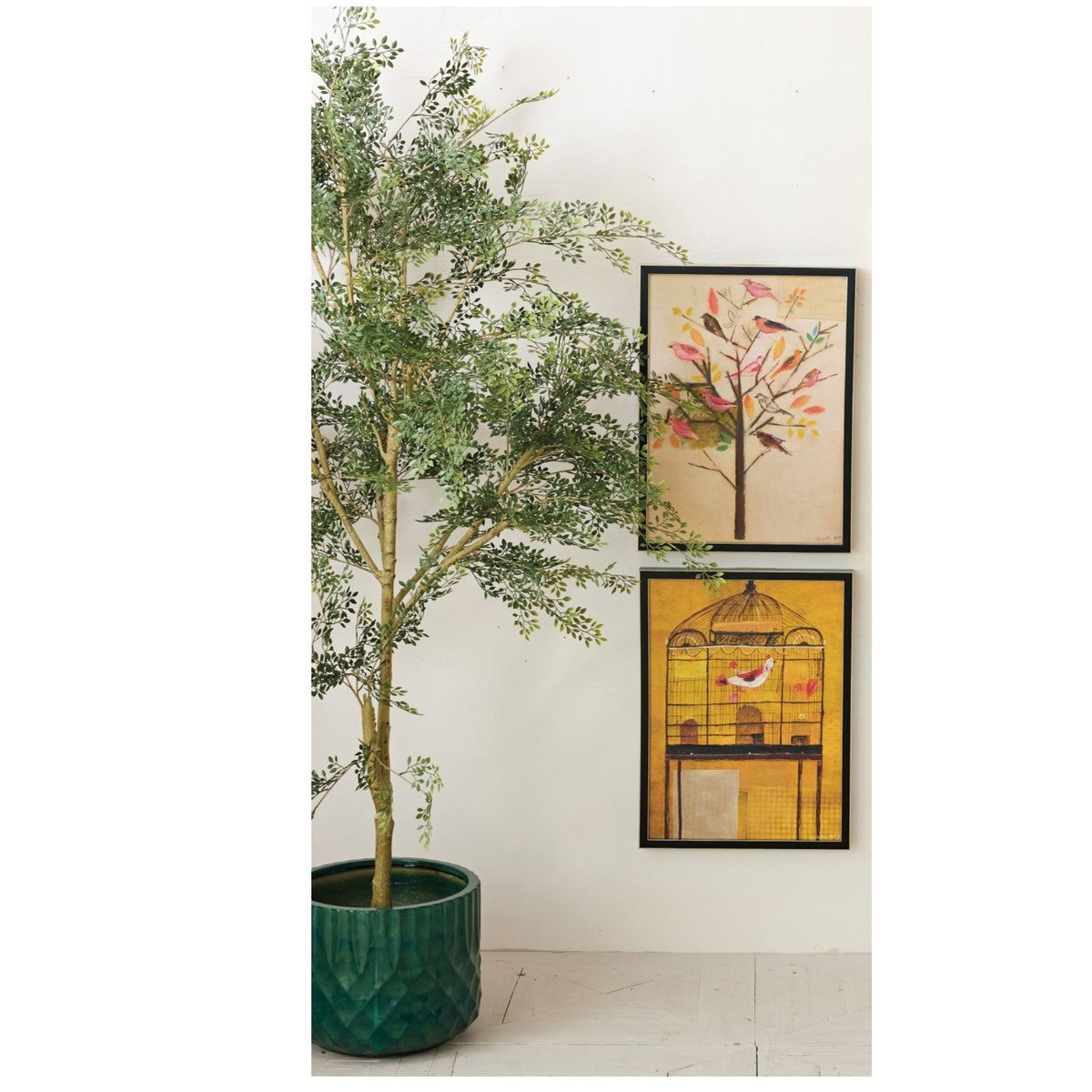 A potted green tree stands beside a white wall decorated with CREATIVE COOP’s FRAMED CANVAS WALL ART WITH HAND-PAINTED BIRDS, featuring a yellow birdcage and perched bird.