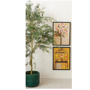 A potted green tree stands beside a white wall decorated with CREATIVE COOP’s FRAMED CANVAS WALL ART WITH HAND-PAINTED BIRDS, featuring a yellow birdcage and perched bird.