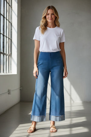 A person stands on a wooden deck with their back to the camera, wearing the FRANK & EILEEN WESTPORT Illusion Cuffed Hem Pull On Jean in 2013 Wash, styled with a white shirt and sandals. A large metal planter filled with greenery sits beside them.