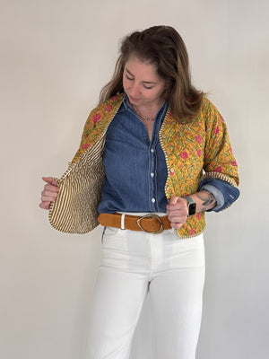 A woman with visible forearm tattoos smiles slightly against a plain background, wearing the FARYLINA ZOIE Reversible Quilted Jacket over a denim shirt, white pants, and a tan belt.