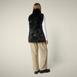 A woman with straight blonde hair wears the SAVE THE DUCK NIVES REVERSIBLE VEST over a white long-sleeve top, beige wide-leg pants, and black shoes, standing against a plain background and highlighting the vest's chic style.