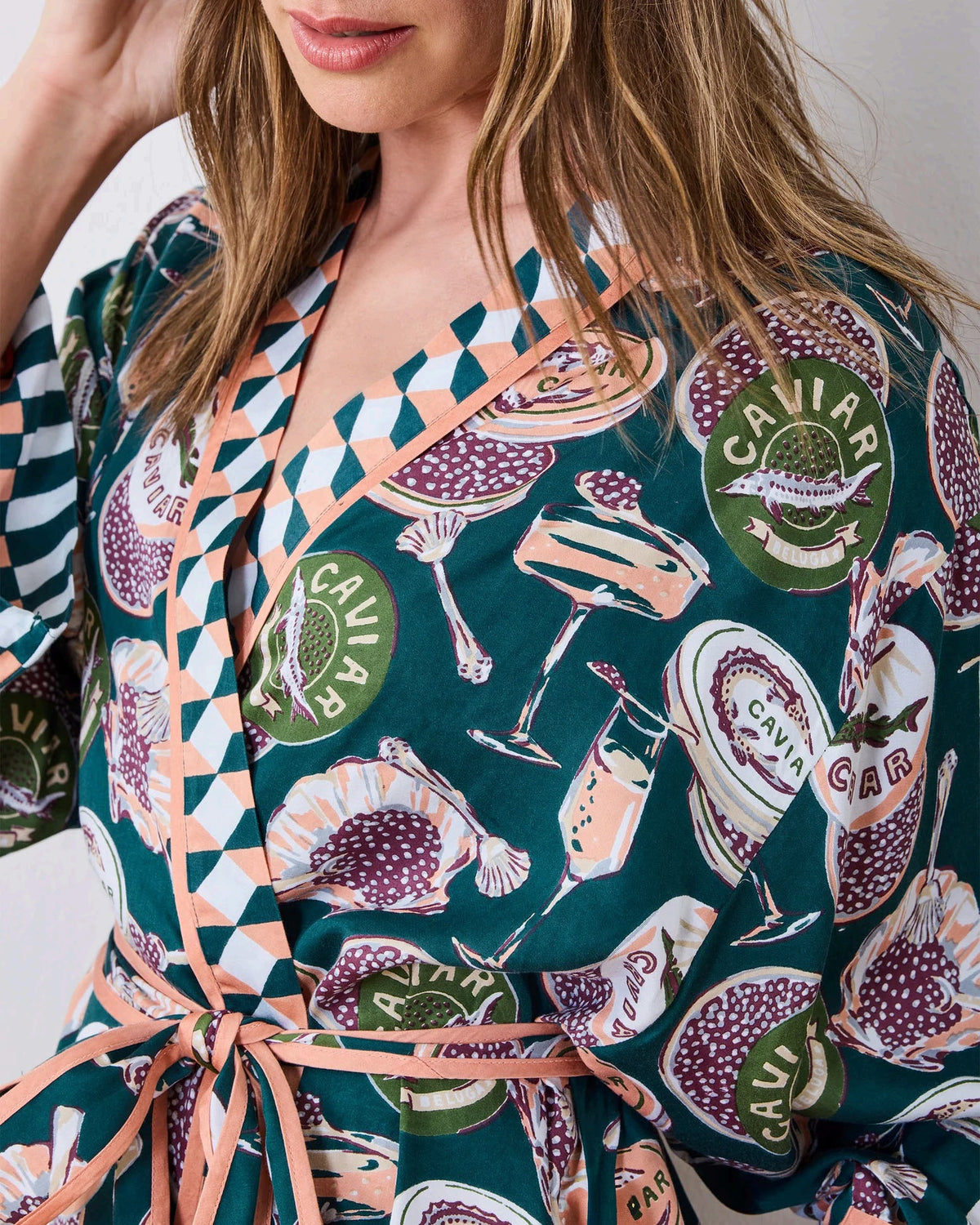 A woman stands barefoot against a white wall, smiling and holding a white cup in one hand as she poses slightly angled to the side. She is wearing the PRINTFRESH Champagne & Caviar Eco Satin Robe in Peacock Feather—a long, colorful eco-friendly robe featuring a vibrant botanical print with peacock feathers and checkered trim.