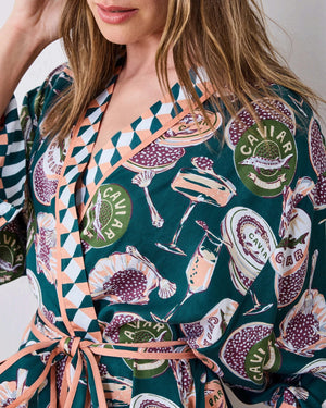 A woman stands barefoot against a white wall, smiling and holding a white cup in one hand as she poses slightly angled to the side. She is wearing the PRINTFRESH Champagne & Caviar Eco Satin Robe in Peacock Feather—a long, colorful eco-friendly robe featuring a vibrant botanical print with peacock feathers and checkered trim.