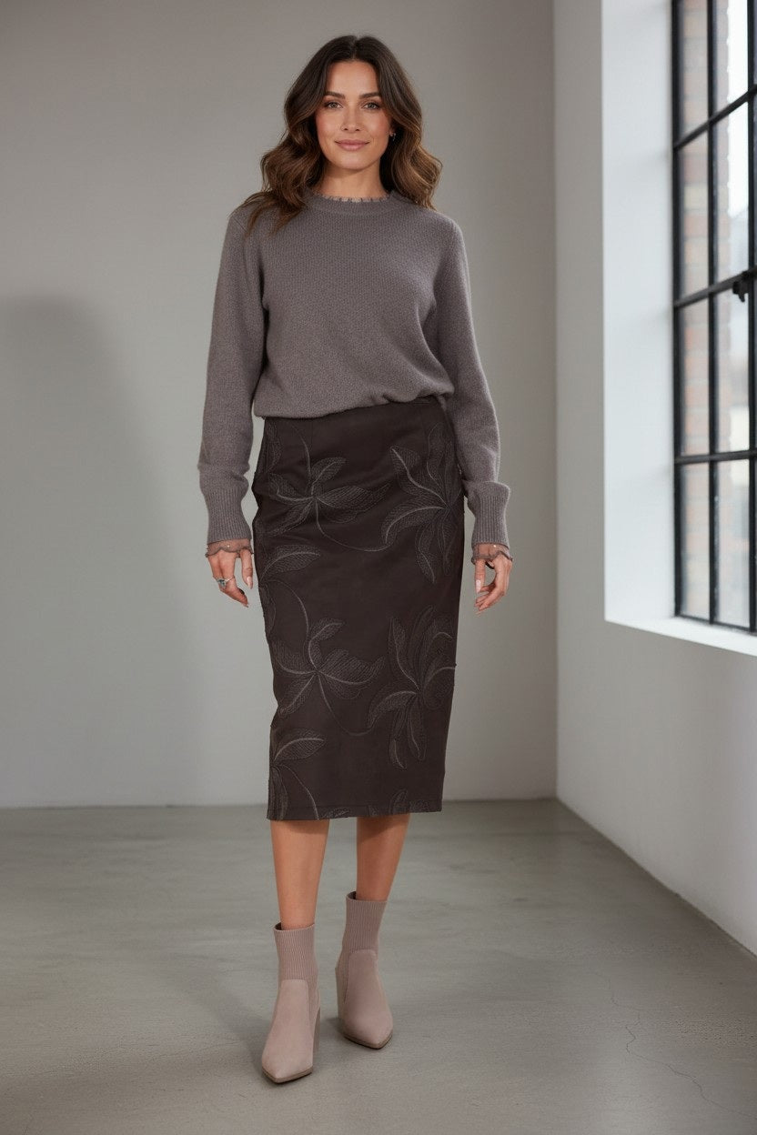 A woman with wavy brown hair wears a gray sweater and the JESSIE LIU MICROSUEDE MIDI SKIRT WITH EMBROIDERY, paired with taupe ankle boots. She stands indoors on a polished floor near large windows.
