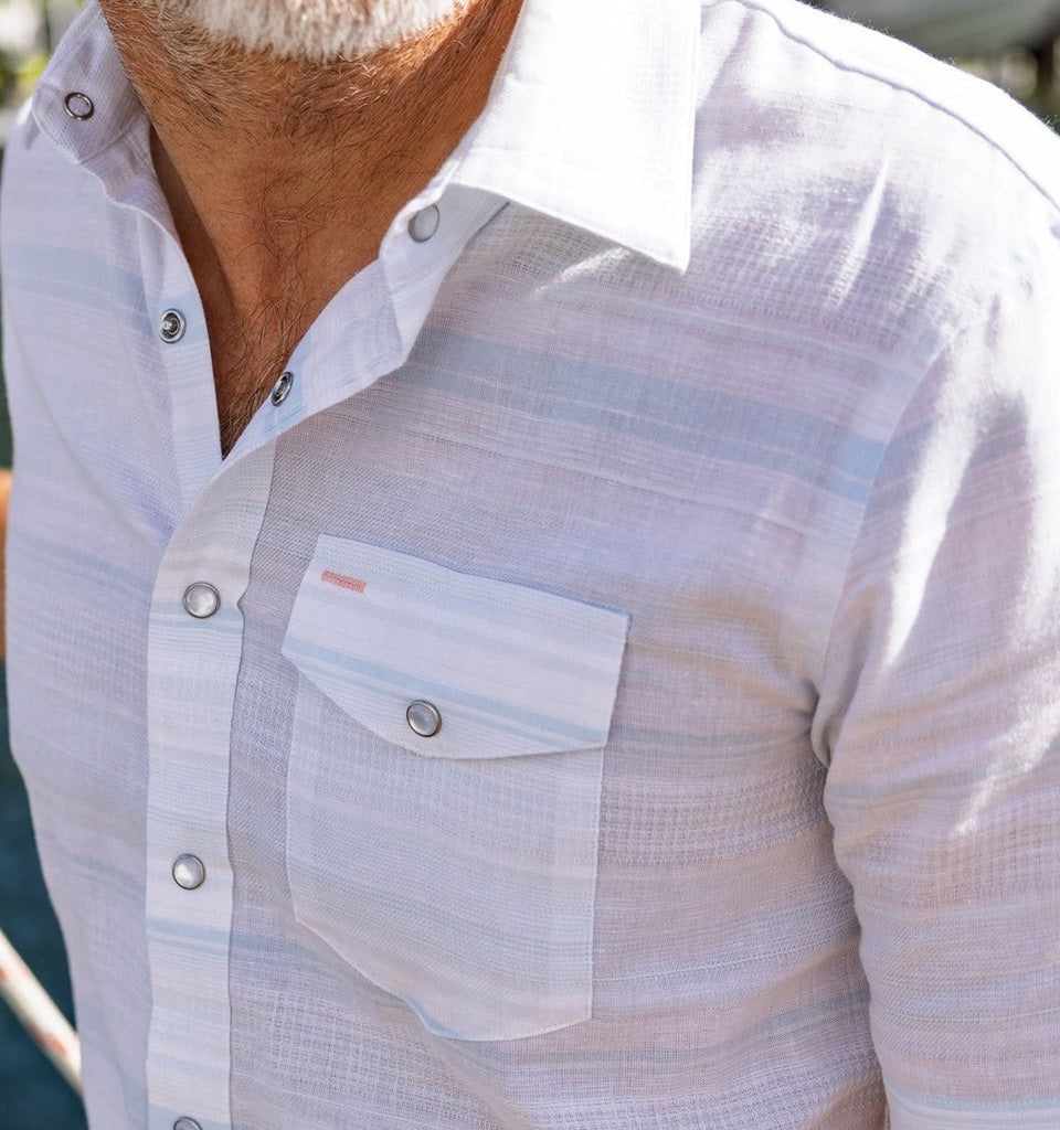 A man wearing the CRIQUET Hemp Stripe Pearl Snap Shirt in Light Blue—a short-sleeve Western style shirt with a chest pocket—smiles and looks to the side.