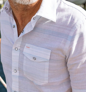 A man wearing the CRIQUET Hemp Stripe Pearl Snap Shirt in Light Blue—a short-sleeve Western style shirt with a chest pocket—smiles and looks to the side.