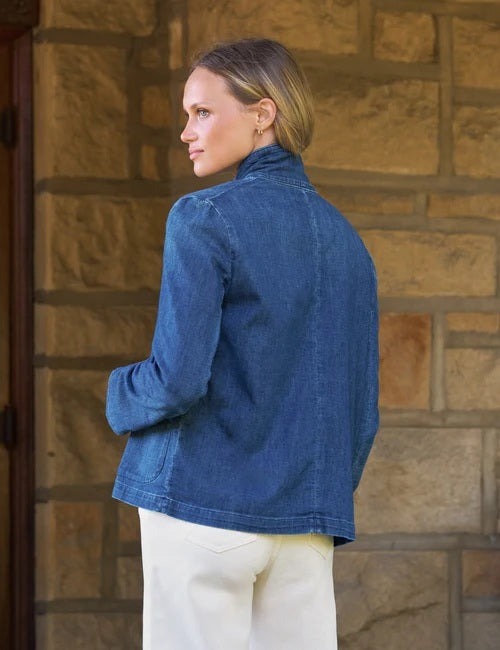 A woman stands smiling in front of a stone wall, wearing the FRANK & EILEEN Mini Belfast Cropped English Peacoat in 1984 Wash Denim over a white top and white pants.