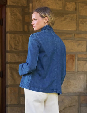 A woman stands smiling in front of a stone wall, wearing the FRANK & EILEEN Mini Belfast Cropped English Peacoat in 1984 Wash Denim over a white top and white pants.