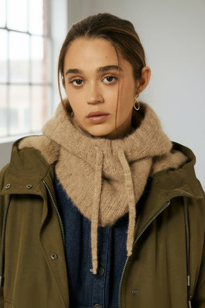 A woman with brown hair pulled back, wearing a green jacket, a navy top, and the HARTFORD - MEVA HOOD IN BEIGE by HARTFORD—a chunky tan knit merino wool scarf—along with a single gold hoop earring, stands against a plain off-white background.