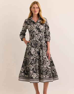 A woman stands wearing the HINSON WU - MARTINA 2/4 SLEEVE PAISLEY PRINT DRESS. This refined cotton dress by HINSON WU features a black background with a white floral and paisley print, a collared neckline, mid-calf length, flared skirt, side pockets, and elegant 2/4 sleeves.