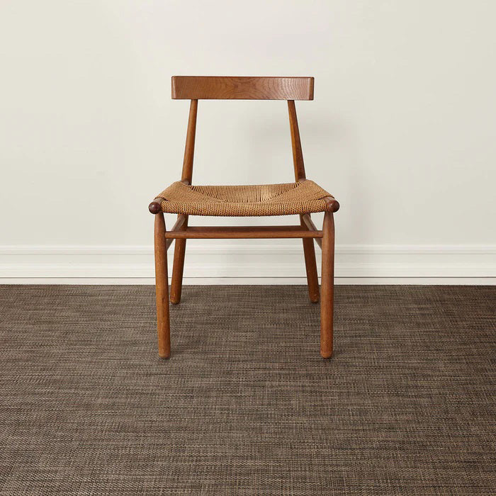 A wooden chair with a woven seat and simple backrest sits on the CHILEWICH - BASKETWEAVE FLOORMAT by CHILEWICH, made with recycled content, set against a plain white wall with white baseboards.
