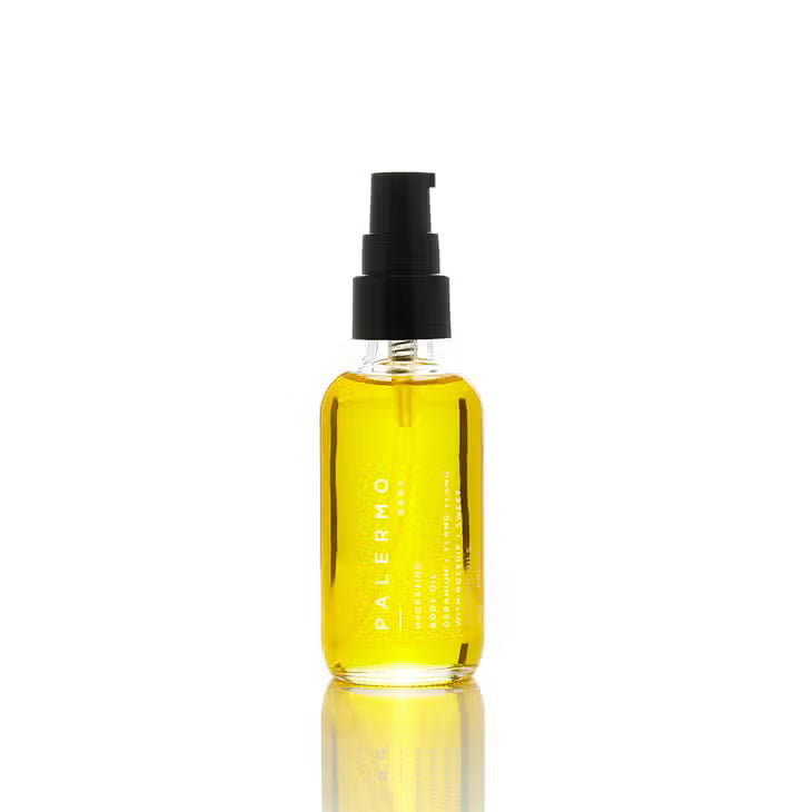 A transparent glass bottle with a pump dispenser, containing yellow liquid infused with restoring oils, labeled "PALERMO - BERGAMOT AND LAVENDER BODY OIL" in white letters. The bottle is isolated on a white background.