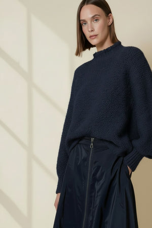 A woman with straight brown hair wears the VICARIO CINQUE MADEIRA Boucle Sweater in midnight blue, paired with a shiny navy skirt, standing against a beige wall dappled with window shadows.
