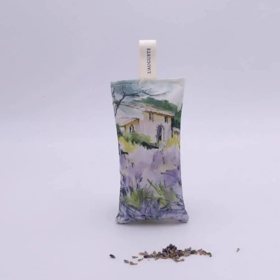 The L&#39;AUGUSTE PROVENCE - FRENCH LAVENDER SACHET FROM PROVENCE features a painted house in a lavender field, stands upright with dried lavender in front, and is topped with a white &quot;L&#39;AUGUSTE&quot; ribbon—authentic from Provence.