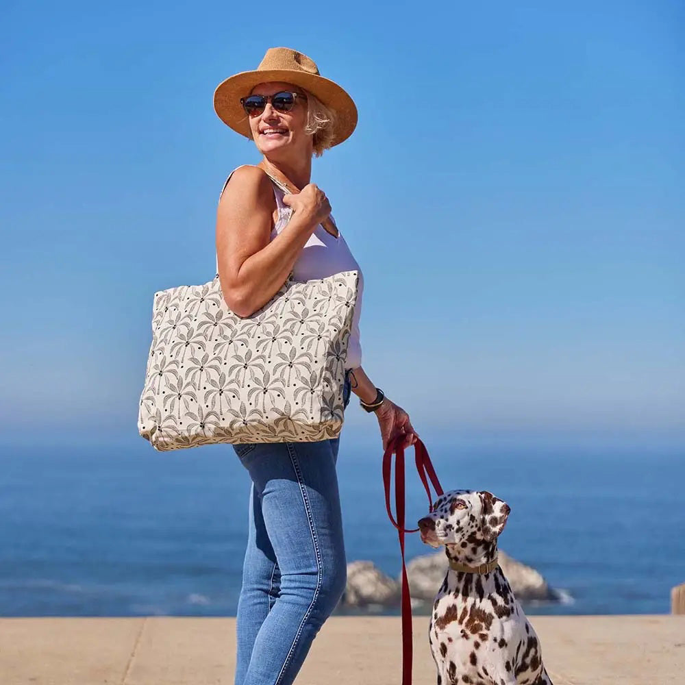 The ROCKFLOWERPAPER PALM SHOPPER TOTE BAG is a spacious cotton canvas bag with two handles, featuring a stylish brown palm tree print on a white background.