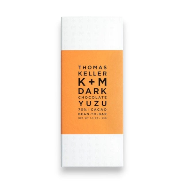 A white chocolate bar wrapper with an orange label reads, “K+M - DARK CHOCOLATE YUZU BAR K & M EXTRAVIRGIN CHOCOLATE 70% CACAO BEAN-TO-BAR NET WT. 1.9 OZ / 55G,” highlighting a distinctive yuzu-flavored dark chocolate made from bean-to-bar Arriba Nacional cacao.