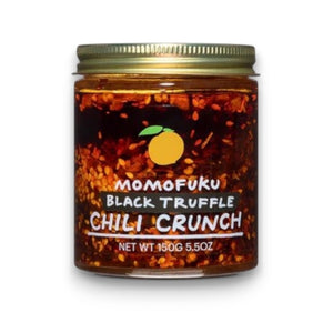 A glass jar of MOMOFUKU - BLACK TRUFFLE CHILI CRUNCH by MOMOFUKU, featuring a gold lid and orange-accented label, filled with chili oil, visible chili flakes, and rich black truffle flavor.