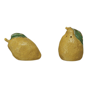 A yellow, lemon-shaped bowl of grains sits on a light surface. In front are CREATIVE COOP STONEWARE LEMON-SHAPED SALT AND PEPPER SHAKERS with green leaves, adding charm to your kitchen décor as grains scatter nearby.