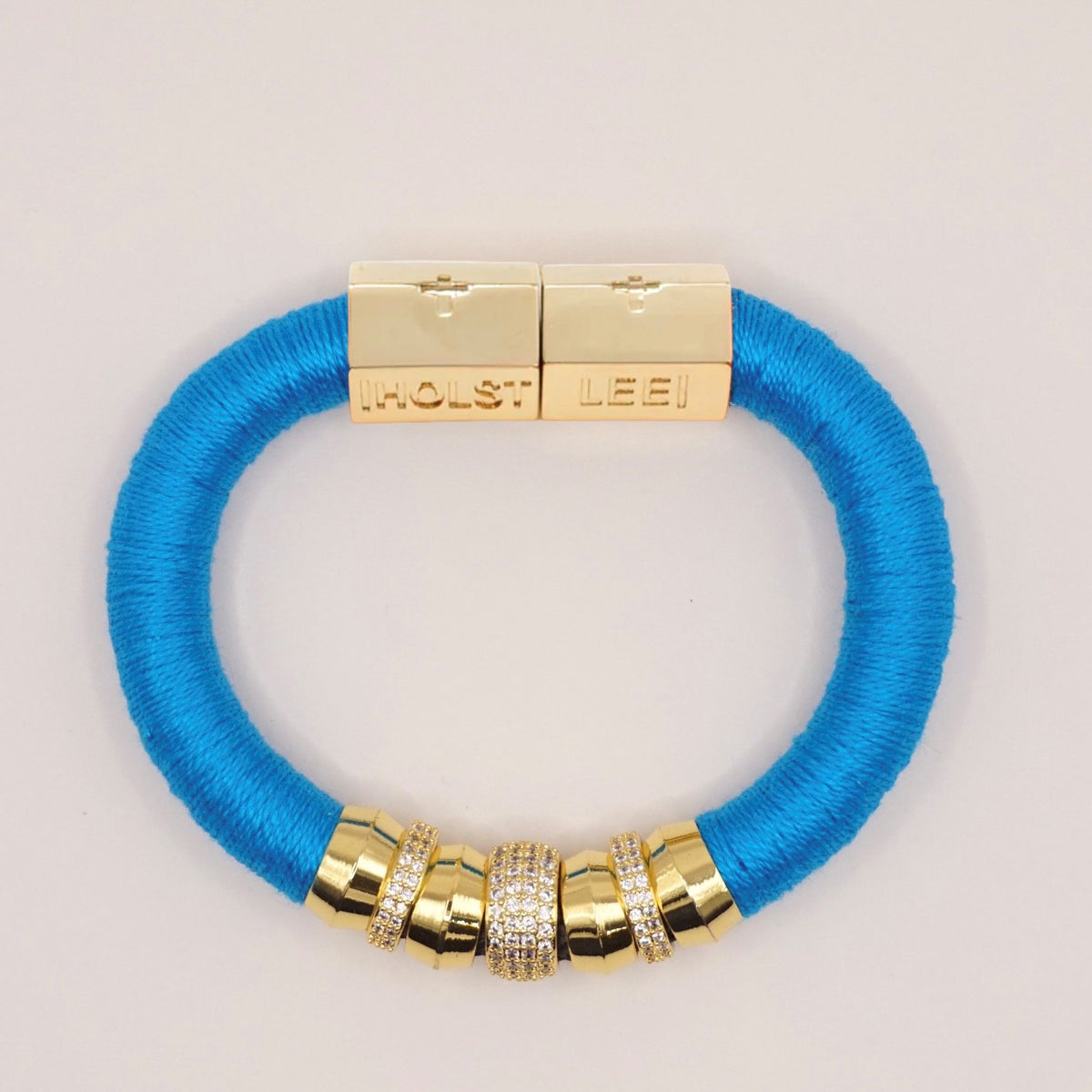 The HOLST + LEE - LUXE CLASSIC BRACELET by HOLST &amp; LEE features an elegant orange cord with gold-plated accents, including a rectangular gold clasp adorned with several gold and diamond-encrusted beads near the clasp.