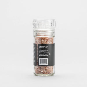 A refillable glass grinder labeled "JACOBSEN SALT - GRINDER PINK HIMALAYAN SALT" from JACOBSEN SALT CO. rests on a light stone surface with a plain gray background, filled with Himalayan pink salt crystals.