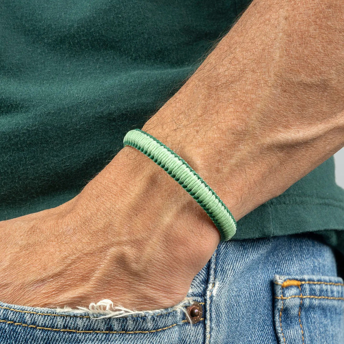 A close-up of a person's wrist wearing the MAKARLA MEN'S REVERSIBLE SURFER BRACELET in mint/hunter green. The person is dressed in a green shirt and blue jeans with a small tear near the pocket.