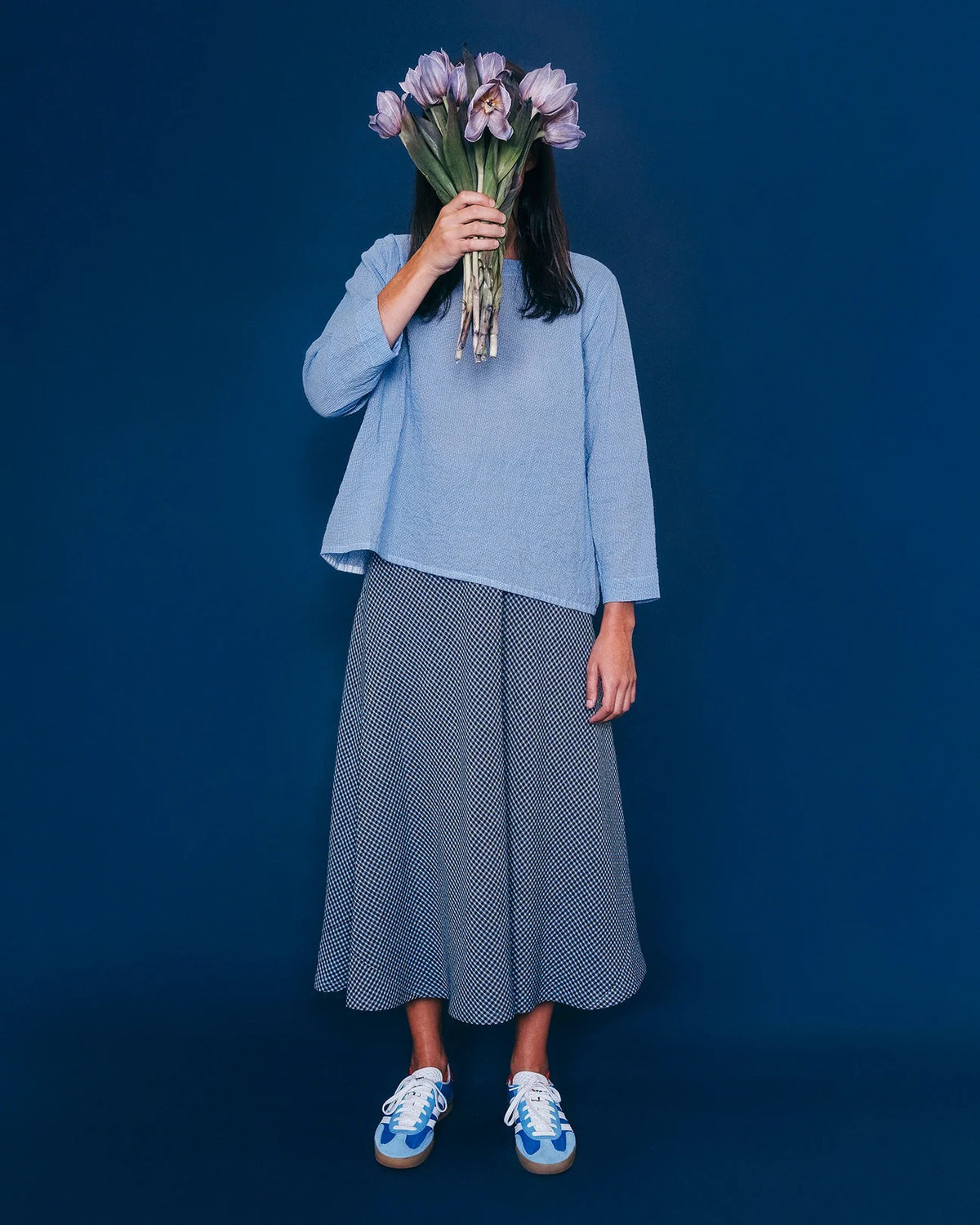 A person stands against a blue background, holding a bouquet of purple flowers in front of their face. They wear a light blue long-sleeve top, a patterned blue skirt, and blue-and-white sneakers.