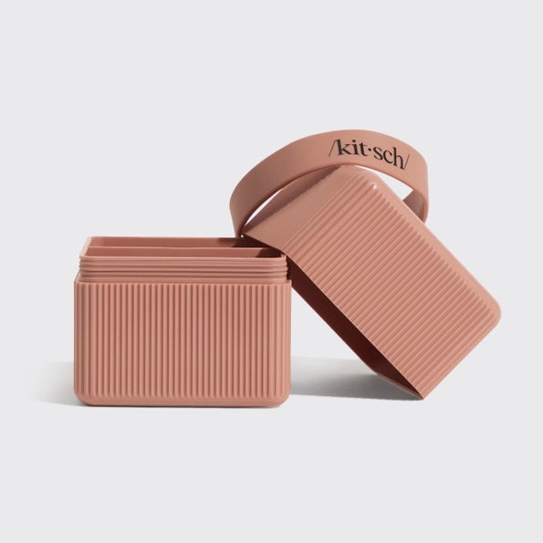 Two ribbed, rectangular KITSCH TWO COMPARTMENT TRAVEL CASES IN TERRACOTTA—one open with its lid tilted to the side—offer a stylish way to organize your beauty essentials and make an ideal travel case. Inside the open compartment are two soap bars, one dark and one yellow, while “kit-sch/” is inscribed on the lid. The background is plain white.
