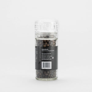 A glass JACOBSEN SALT - GRINDER TELLICHERRY PEPPERCORNS jar from JACOBSEN SALT CO. rests on a light stone surface with a plain gray background, highlighting this premium pre-filled grinder.